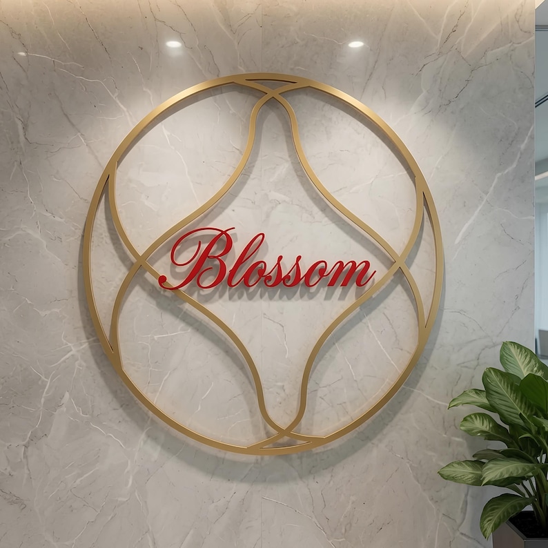 May include: A circular gold metal wall sign featuring an intricate, symmetrical geometric frame. The word Blossom is written in a red, cursive script font across the center of the frame. The sign is mounted on a light gray marble wall surface.