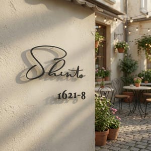 May include: A black metal wall sign featuring the word Shinto in a cursive script font, positioned above the numbers 1621-8 in a standard sans-serif font. The sign is mounted on a light-colored, textured stucco wall surface.