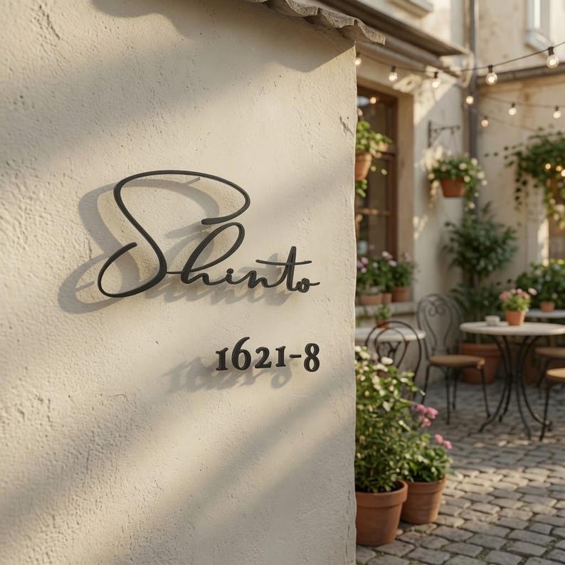 May include: A black metal wall sign featuring the word Shinto in a cursive script font, positioned above the numbers 1621-8 in a standard sans-serif font. The sign is mounted on a light-colored, textured stucco wall surface.