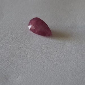 以下が含まれることがあります： ファセット加工された表面を持つ、深いマゼンタピンク色の洋ナシ型の宝石。尖った端と丸みを帯びた基部が特徴で、半透明の質感があり、内部のテクスチャと光を反射する幾何学的なファセットがはっきりと見えます。
