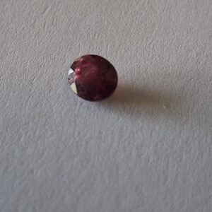 Könnte beinhalten: Ein kleiner, runder, facettierter Edelstein mit einem tiefen rot-violetten Farbton. Der Stein hat eine polierte Oberfläche mit mehreren geometrischen Schliffen, die das Licht reflektieren und subtile Glanzlichter und Schatten auf dem runden Körper erzeugen.