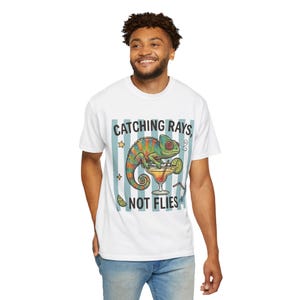 May include: A white short-sleeve t-shirt featuring a graphic of a colorful chameleon sitting on a cocktail glass. The text reads CATCHING RAYS, NOT FLIES. The design includes light blue vertical stripes, a lime wedge, and small star icons.