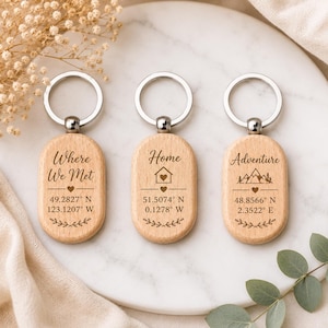 May include: Three oval-shaped light wood keychains with silver metal rings. Each keychain features laser-engraved text and symbols: 'Where We Met' with coordinates, 'Home' with a house icon and coordinates, and 'Adventure' with mountain icons and coordinates.