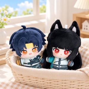 May include: Two plush dolls with embroidered facial features. The left doll has dark blue hair, a yellow headband, and orange eyes. The right doll has black hair, pointed animal ears with white accents, and red eyes. Both wear detailed outfits.
