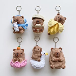 May include: Six crocheted capybara keychains. One wears blue headphones and a scarf holding a white rabbit. One sits in a beige cup. One rests inside a yellow banana peel. One sits in a pink lotus flower. One rides a white goose. One is plain.