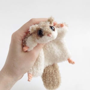 May include: A small, crocheted flying squirrel plush toy with fuzzy light brown and cream-colored yarn. It features large, round, glossy black eyes, a tiny pink nose, and small pink-colored paws. The creature has a flat, extended body shape.