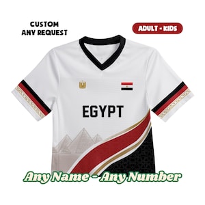 May include: A white V-neck jersey featuring the word EGYPT in black letters, the Egyptian flag, and a gold emblem. The design includes a graphic of pyramids, a diagonal red and gold stripe, and black patterned sections on the sleeves and hem.