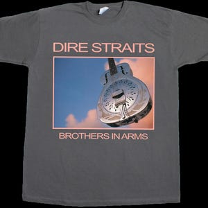 Puede incluir: Una camiseta gris carbón de manga corta con un estampado gráfico de una guitarra resonadora metálica sobre un cielo azul y naranja. El texto DIRE STRAITS aparece sobre la imagen y BROTHERS IN ARMS está impreso debajo en letras rosa claro.
