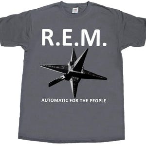 Puede incluir: Una camiseta de manga corta de color gris carbón con el texto blanco R.E.M. en la parte superior, un gráfico en blanco y negro de un objeto con forma de estrella de cinco puntas en el centro y el texto blanco AUTOMATIC FOR THE PEOPLE abajo.