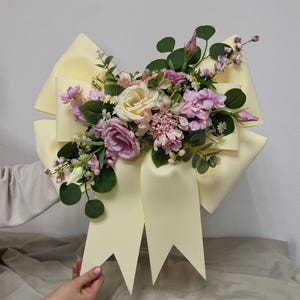 May include: A large, pale yellow decorative bow made of smooth material, adorned with an arrangement of artificial flowers in shades of cream, soft pink, and lavender, accented with green eucalyptus leaves and small white daisy-like blossoms.