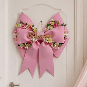 May include: A large pink decorative bow hanging on a white door. The bow is embellished with a variety of small artificial flowers in shades of pink, yellow, and white. A gold-coloured script nameplate reading "Amelia" is centred on the bow.