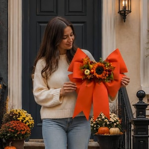 May include: A large, vibrant orange fabric bow featuring a central decorative arrangement of a sunflower, small pumpkins, and autumn foliage. The bow has long, flowing tails and is designed for seasonal door or wall decor, showcasing a rich fall color palette.