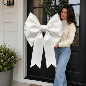 May include: A large, oversized white decorative bow with a smooth, satin-like finish. The bow features two prominent, voluminous loops and two long, wide tails with V-shaped notched ends, creating a structured and symmetrical appearance.