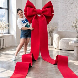 Puede incluir: Un lazo decorativo rojo de gran tamaño hecho de un material similar al fieltro. El lazo presenta un nudo central con dos bucles voluminosos y dos cintas largas y anchas que se extienden hacia abajo y se posan en el suelo con extremos en V.
