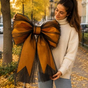 Puede incluir: Un gran lazo decorativo confeccionado con capas de tul negro transparente y tela de color bronce metálico. El lazo presenta un nudo central y dos cintas largas y anchas con extremos puntiagudos, mostrando una paleta de colores negro y bronce.