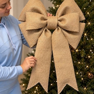 May include: A large decorative bow crafted from tan burlap fabric. The bow features a central knot with two wide, structured loops on each side and two long, downward-hanging tails with V-shaped notched ends, showcasing a coarse, woven texture.