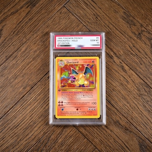 Könnte beinhalten: Eine 1999 1st Edition französische Charizard holografische Sammelkarte in einer durchsichtigen Schutzhülle. Das Etikett oben zeigt 1999 POKEMON FRENCH DRACAUFEU - HOLO 1ST EDITION GEM MT 10. Die Karte zeigt eine Illustration eines Feuerdrachen.