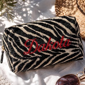 May include: A rectangular zippered pouch featuring a black and cream zebra stripe pattern with the name Dakota embroidered in red script on the front. The pouch has a gold-toned zipper pull and a textured fabric construction.