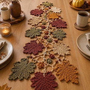 Könnte beinhalten: Ein gehäkelter Tischläufer mit einem zentralen Rankenmuster, das mit verschiedenen herbstfarbenen Blättern in den Tönen Dunkelrot, gebranntes Orange, Olivgrün und Creme verziert ist. Das handgefertigte Textil zeigt ein wiederkehrendes Blattdesign.