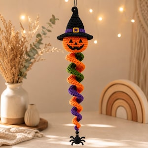 Peut inclure: Une décoration suspendue d'Halloween au crochet représentant une tête de citrouille orange portant un chapeau de sorcière noir avec une bande violette et une boucle jaune. Sous la tête, une longue queue en spirale orange, verte et violette se termine par une petite araignée noire.