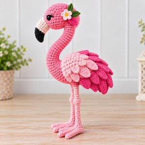 Può includere: Un giocattolo a forma di fenicottero realizzato all'uncinetto in varie tonalità di rosa, con un lungo collo ricurvo, un becco con la punta nera e un piccolo fiore bianco con foglie verdi applicato sul lato della testa. Il corpo ha ali a strati.