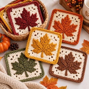 May include: A collection of square crochet coasters featuring raised maple leaf motifs in autumn colours including deep red, orange, yellow, olive green, and brown, each centred on a cream-coloured square base with a decorative openwork border.