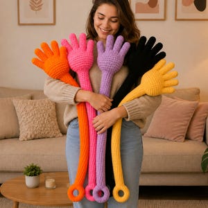 Puede incluir: Un conjunto de cinco peluches largos tejidos a ganchillo con forma de mano en colores naranja brillante, rosa intenso, lavanda, negro y amarillo. Cada peluche presenta una mano de cinco dedos arriba y un anillo circular abajo, con textura.