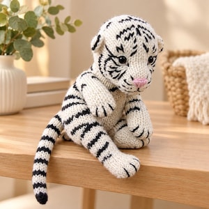May include: A hand-knitted white tiger plush toy featuring black stripes, a soft pink nose, and dark round eyes. The stuffed animal is crafted from yarn with a visible knit texture, showing a seated posture with a long tail extending to the side.