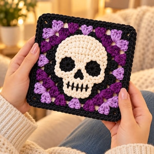 Puede incluir: Un cuadrado de ganchillo tipo granny square que presenta un motivo central de calavera en color blanco roto con cuencas oculares negras y dientes bordados. La calavera está rodeada por capas de hilo negro, morado oscuro y lavanda claro.