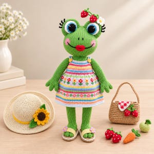 May include: A green crocheted frog doll wearing a multi-colored striped dress with floral patterns, red lips, and strawberry hair accessories. The set includes a straw hat with a sunflower, a small woven bag, and miniature crocheted food items.