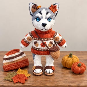 May include: A knitted husky plush toy wearing an orange and white sweater with acorn and leaf patterns, a matching acorn-shaped crossbody bag, and sandals. Beside it are a striped knit beanie, two crocheted pumpkins, and several fabric leaves.