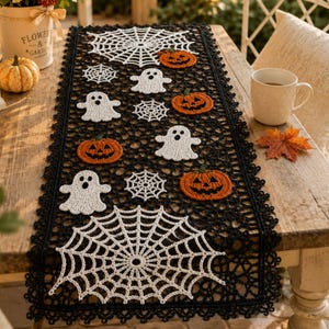 May include: A black crocheted table runner featuring white spider web motifs, white ghost figures, and orange jack-o'-lantern pumpkin designs. The runner has a scalloped lace-like border and is laid out flat on a wooden surface.