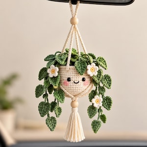 May include: A crocheted hanging plant ornament featuring a cream-colored pot with a smiling face, pink cheeks, and black eyes. The pot contains green crocheted leaves and small white flowers with yellow centers, suspended by cream cords with wooden beads.