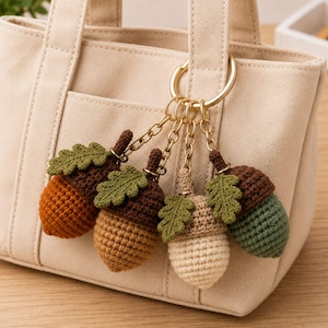 May include: A gold-toned metal keyring featuring four crocheted acorn charms, each with a textured green leaf attached. The acorns are crafted in varying shades of brown, beige, and green yarn, suspended from individual chains on a circular ring.