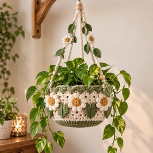 Pode incluir: Um suporte de plantas em crochê nas cores creme e verde sálvia, com motivos de margaridas brancas tridimensionais com centros amarelos e detalhes de folhas verdes. Inclui contas de madeira natural e uma planta pothos verde.