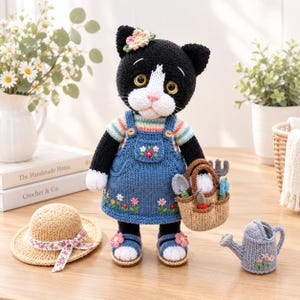 以下が含まれることがあります： 黒と白の編みぐるみの猫の人形。花柄の刺繍が施された青いニットのオーバーオールスカート、ストライプのシャツ、花で飾られたサンダルを着用しています。猫はミニチュアのガーデニングツールが入った小さな編みかごを持ち、麦わら帽子の横に立っています。