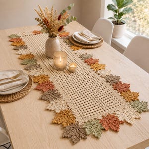 May include: A rectangular crochet table runner featuring a central grid pattern bordered by a series of crocheted maple leaves in shades of cream, tan, mustard yellow, sage green, and burnt orange, arranged along the edges of the runner.