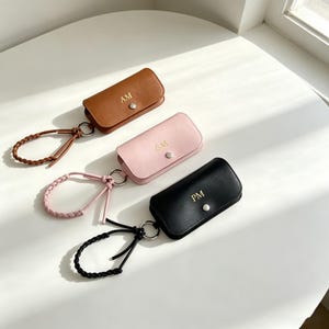 May include: Three rectangular key cases in brown, light pink, and black, each featuring a silver snap button closure, a matching braided wrist strap attached to a silver ring, and gold embossed initials reading AM, GM, and PM respectively.