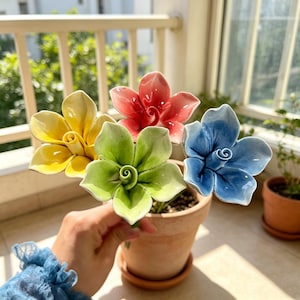 May include: A collection of four ceramic flowers in yellow, red, light green, and blue, each featuring five smooth, curved petals and a central spiral detail. The flowers are arranged together, showcasing a glossy finish and vibrant colors.
