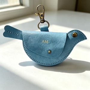 May include: A light blue bird-shaped pouch with a textured finish, featuring the gold-stamped letters AM on the front flap. The pouch includes a brass-toned metal snap closure and a matching swivel lobster clasp attached to the top for hanging.