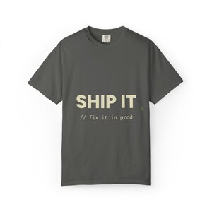 以下が含まれることがあります： ダークグレーの半袖Tシャツ。クリーム色の大きな文字で「SHIP IT」、その下に小さな文字で「// fix it in prod」とプリントされています。クルーネックの標準的なフィット感で、テキストの近くに小さな緑色のアイコンが配置されています。