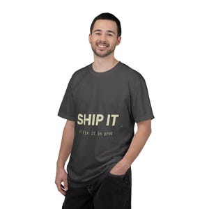 May include: A dark gray short-sleeve t-shirt featuring the text "SHIP IT" in large light yellow letters, with the smaller text "//fix it in prod" printed underneath in a monospaced font, and a small green icon located to the right.