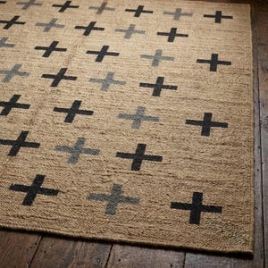 May include: A rectangular area rug featuring a natural tan woven base with a repeating grid pattern of dark charcoal and light grey plus signs. The rug has a textured, fibrous surface and is laid out flat, displaying a consistent geometric design.