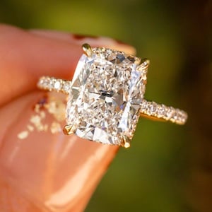 Puede incluir: Un anillo de oro con una gran piedra central rectangular de corte cojín con bordes facetados, sujeta por cuatro garras. La banda delgada está incrustada con una hilera de pequeñas piedras redondas y transparentes en la mitad superior.