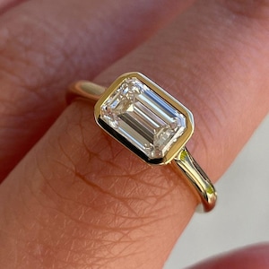 Può includere: Un anello in oro con una pietra preziosa trasparente a taglio smeraldo rettangolare montata in una lunetta liscia. La fascia è in oro giallo lucido con un profilo arrotondato, che mette in risalto la pietra sfaccettata al centro.