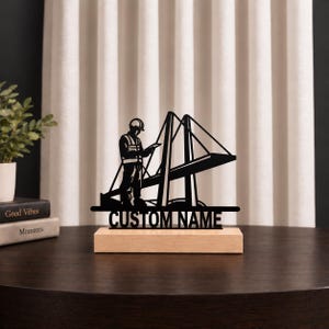 May include: A black metal silhouette of a construction worker wearing a hard hat and safety vest, standing next to a bridge structure. The design is mounted on a rectangular light-colored wooden base with the text CUSTOM NAME at the bottom.