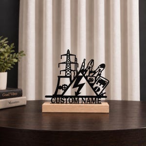 May include: A black metal silhouette display piece featuring an electrical transmission tower, a lightning bolt, and various tools, mounted on a rectangular light wood base. The text CUSTOM NAME is cut out in capital letters at the bottom.