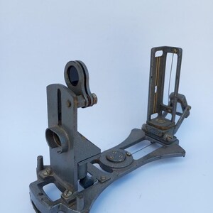 May include: A vintage gray metal surveying instrument featuring a vertical sight with a circular lens, a rear vertical frame with a thin wire, and a central circular dial. The device has a curved base with adjustable screws and mechanical components.