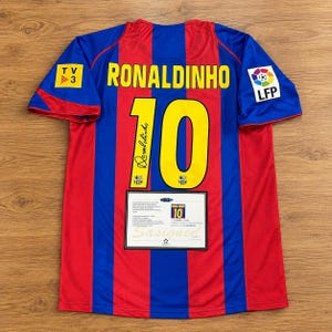 May include: A red and blue striped football shirt featuring the name RONALDINHO and the number 10 in yellow on the back. The shirt includes a TV3 logo, an LFP patch, a signature on the number 10, and a certificate of authenticity label.