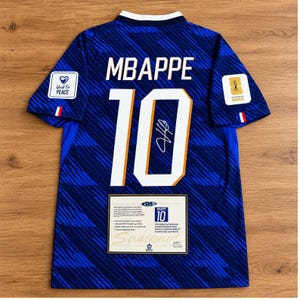 Puede incluir: Una camiseta de fútbol azul oscuro con el nombre MBAPPE y el número 10 en blanco junto a una firma negra. Las mangas tienen parches que dicen World For PEACE y FIFA WORLD CUP 2022. Se incluye un certificado de autenticidad rectangular.